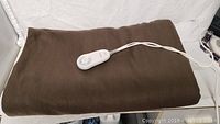 Full view of folded brown heated throw blanket with attached power cord and controller on white surface.
