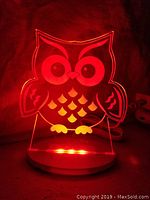 Lit owl-shaped acrylic LED night light with red light glowing on circular white base.