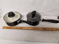 Front view of two black non-stick sauce pans with glass lids and black handles aligned next to a ruler showing their length.