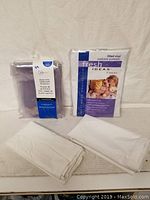 Four items: one tablecloth protector in packaging, one packed mattress protector, two folded white pillow covers on white surface.