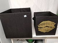 Two black fabric cube storage bins, one featuring a gold lip print design on the front, both square shaped with open tops and handles on the sides.