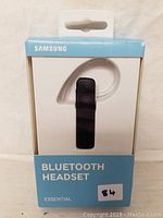 Front view of packaged Samsung Bluetooth Headset Essential in white and blue box, showing the headset with black body and clear ear hook.