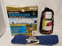 Three items: ADEQ windshield cover box, jumper cables in black carrying bag, and blue microfiber shop towels in plastic wrapping.