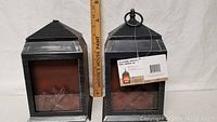 Two black distressed metal LED glowing fireplace lanterns side by side with wood ruler showing height.