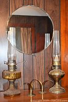 Two vintage hurricane lamps, one amber glass with ribbed base, one brass with intact fuel filter, in front of round wall mirror on wood panel wall, alongside two brass candle holders.
