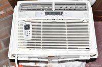 Frigidaire white window air conditioner with remote lying on top of the unit showing front and ventilation slats.