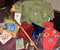 Full view of Boy Scout uniforms, merit badge sash, kerchiefs, patches, wooden bow, and assorted scouting items piled on a chair and table.