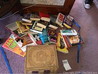 Image showing antique Webster American Dictionary with various paperback books including mystery novels and cat books scattered around on carpet.