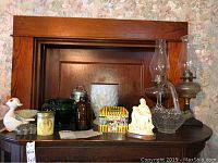 Photo of the full collection arranged on dark wooden furniture showing two hurricane lamps, various glass and ceramic pieces including milk glass items, religious figurines, and novelty collectibles.