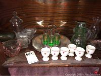 Photo showing collection of glassware including green Vaseline shot glasses on a metal stand, white egg cups, pink and clear depression glass bowls, and decanters on a wood shelf.