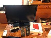 LG 19 inch TV shown with digital alarm clock, two remotes, and boxed red Bose SoundLink Color Bluetooth speaker on wooden table.