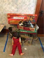 Vintage doll sitting on carpeted floor wearing red overalls and beige shirt, next to various vintage toys and boxed games