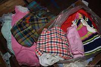 Assorted kids vintage clothing shown in a cardboard box with various colors and materials visible including pinks, plaids, and stripes.