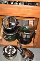 Pots and pans stacked inside a wooden cabinet including black pots, green pot, and stainless steel pot with glass lids.