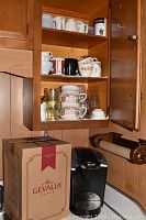 Kitchen cabinet with assorted mugs and glassware and Gevalia boxed coffee maker on counter beneath