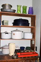 Wooden shelving unit holding multiple vintage kitchen items including enamelware cooking pots, metal pans, muffin tins, and glass vases.