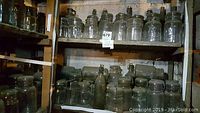 Multiple shelves filled with glass jars and bottles of different sizes, most clear or slightly tinted glass. Many jars have wire bail lids or metal lids. Items dusty from long storage in basement.