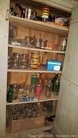 Open wooden cabinet with multiple shelves showing assorted glass jars, bottles, ceramic mugs, and kitchen containers
