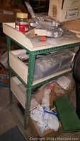 Side view of green metal workshop shelf with wooden drawers and various tools and hardware on and inside it.