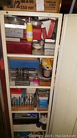 Interior of white metal cabinet with multiple shelves containing red and grey toolboxes, plastic bins and a yellow glue applicator bottle