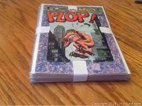 Photo of the top and side view of the stack of 10 comics banded together, showing the cover of one comic titled PLOP! with cartoon artwork