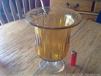 Full view of the amber hand blown glass vase showing its flared rim and pedestal base with a red lighter for scale on a wooden table.