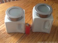 Photo showing two white milk glass canisters with silver lids each on a wooden table next to a red lighter for size reference.