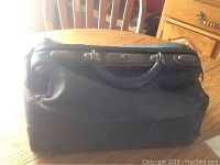 Front and top side of the vintage black doctor's bag showing handle, metal clasps, and wear on edges.