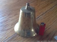 Photo of old brass bell on wooden floor next to a red lighter for size reference.