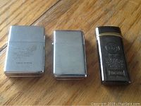 Three lighters placed upright on wooden surface: two metal Zippo-style (one engraved with Nova Scotia), one black and gold windproof lighter.