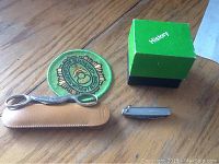 Green embroidered patch, small magnifier in leather case, small pocket knife, and green cube with 'History' label arranged on wooden floor.