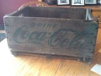 Full view of antique wooden Coca Cola crate on wooden floor showing the front with faint green Coca Cola logo and drink text.