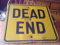 Full view of the yellow metal Dead End sign with black lettering showing wear and rust spots.