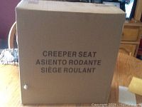 Closed intact box labeled 'Creeper Seat Asiento Rodante Siège Roulant' in multiple languages, showing packaging condition.
