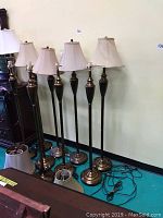 Nine brass tone floor lamps standing side by side with some shades on them, against a pale wall, cords visible on floor.