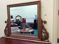 Full frontal view of large standing rectangular beveled mirror with gilded frame and decorative side scroll elements on wooden base shelf.