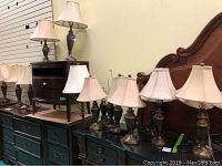 Several metal base table lamps with fabric shades displayed on wooden furniture.