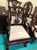 Two wooden arm chairs featuring carved backs with ornate floral motifs, cream fabric upholstered seats with damask pattern, and claw paw feet.