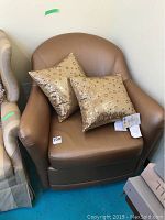 Brown leather chair with two gold decorative pillows sitting on it, showing the full chair and pillows from a frontal angle.