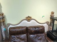 Full view of white upholstered headboard with carved wooden frame and gold accents behind brown chairs.