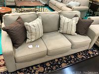 Full front angle view showing the beige upholstered couch with three seat cushions and four decorative pillows, placed on a patterned area rug.