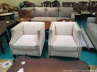 Front view of two upholstered chairs with grid-patterned beige fabric, placed in a showroom with other furniture items around.