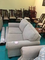 Front and side angle view of light gray upholstered loveseat with cushions and rolled arms surrounded by other wooden chairs.
