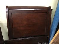 Photo of a dark stained wooden headboard with rectangular panel and framed trim, showing the top and side edges with some surface scratches.