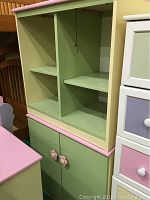 Front view of light green and pink child's cubby cabinet with two open cubbies above and double-door storage below with flower handles