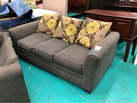 Front view of three-seat gray upholstered sofa with floral pillows on showroom floor.