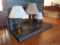 Three table lamps on a fireplace hearth, two tall brass lamps with white pleated shades, one smaller lamp with a white conical shade, showing overall condition and size relation