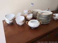 Photo showing 9 cups, plates stacked in two piles, sugar bowl, creamer, and a covered dish, all decorated with floral patterns and gold trim.