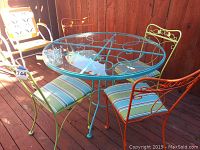 Glass top patio table with three colorful metal chairs around it on a wooden deck, showing overall appearance and size relative to chairs.