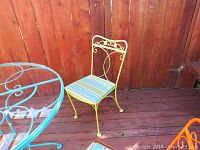 Yellow metal patio chair with striped blue, green, white cushion, decorative back design, wooden fence background